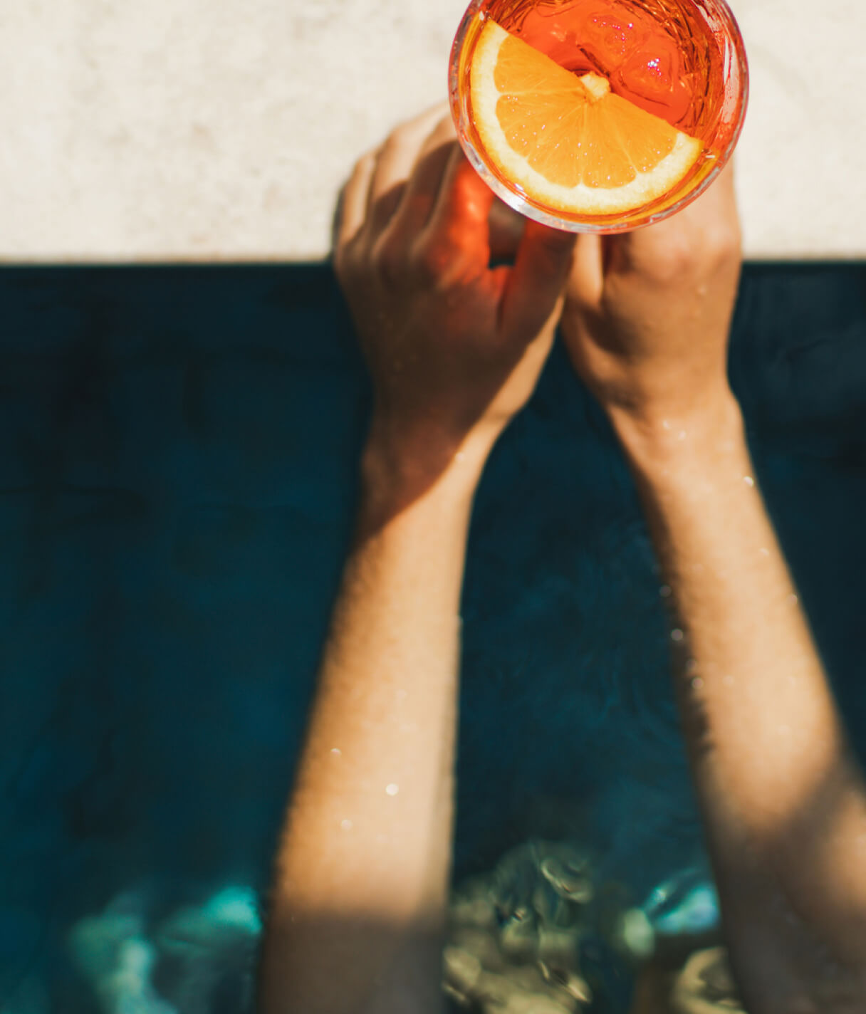 A woman’s hands holding a vibrant orange drink at the poolside, sunlight bounces across the bright blue pool water